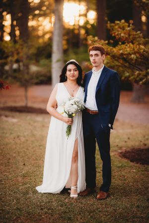 A couple poses together outdoors at sunset in wedding attire with fall foliage.