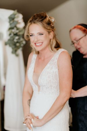 A bride wearing a white lace wedding dress smiles warmly while getting ready for her wedding ceremony.