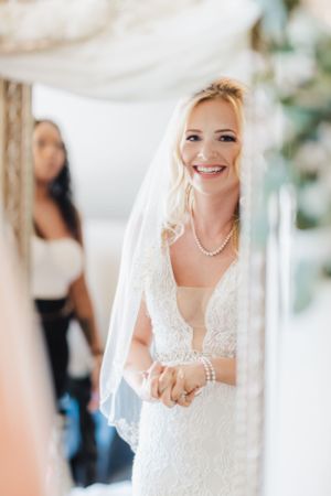 A bride wearing a beautiful white lace wedding dress with pearls and veil smiles while getting ready.