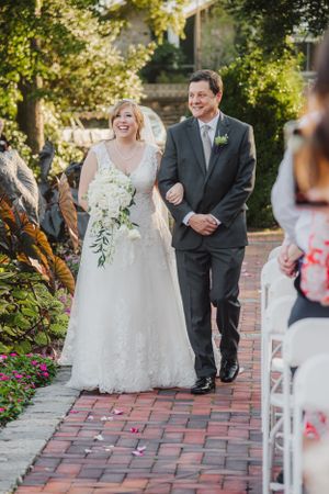 Wedding couple shares joyful moment walking down brick pathway lined with flowers.