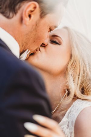 A romantic close-up of a tender kiss between newlyweds, with the bride in white lace and pearls and groom in navy suit.