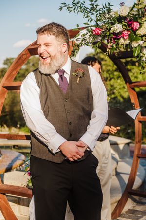 Groomsman in brown vest and tie laughs during outdoor garden ceremony.