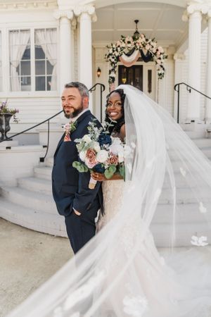 A wedding couple poses on white mansion steps under a floral arch.