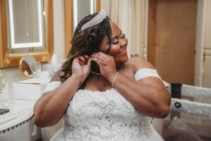 Person putting on earrings while wearing white wedding dress in sunlit room.