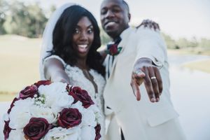 Joyful wedding couple showing off ring and bouquet outdoors by lake.