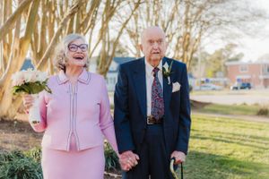 Senior couple holding hands outdoors, wearing pink and navy formal attire.