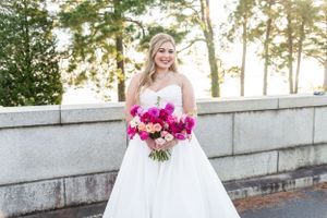 Bride in white wedding gown holds vibrant pink bouquet while posing against stone wall at sunset.