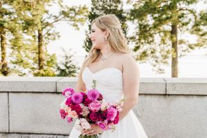 A bride in a white dress holds a vibrant pink and magenta bouquet against a sunny outdoor backdrop.