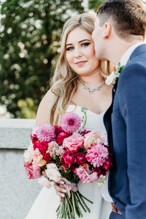 A romantic wedding portrait of a couple with pink flowers and greenery in an outdoor setting.