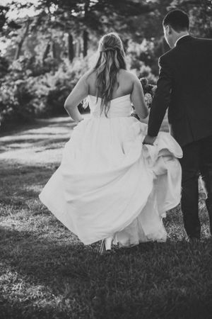 Black and white photo of newlyweds walking together on a tree-lined path.