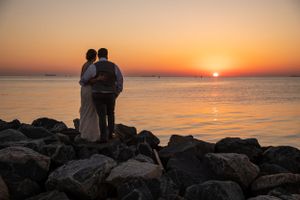Silhouetted couple standing on rocky shore watching vibrant orange sunset over calm ocean waters.