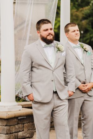 Groomsmen in light gray suits stand at a white pergola during outdoor ceremony.
