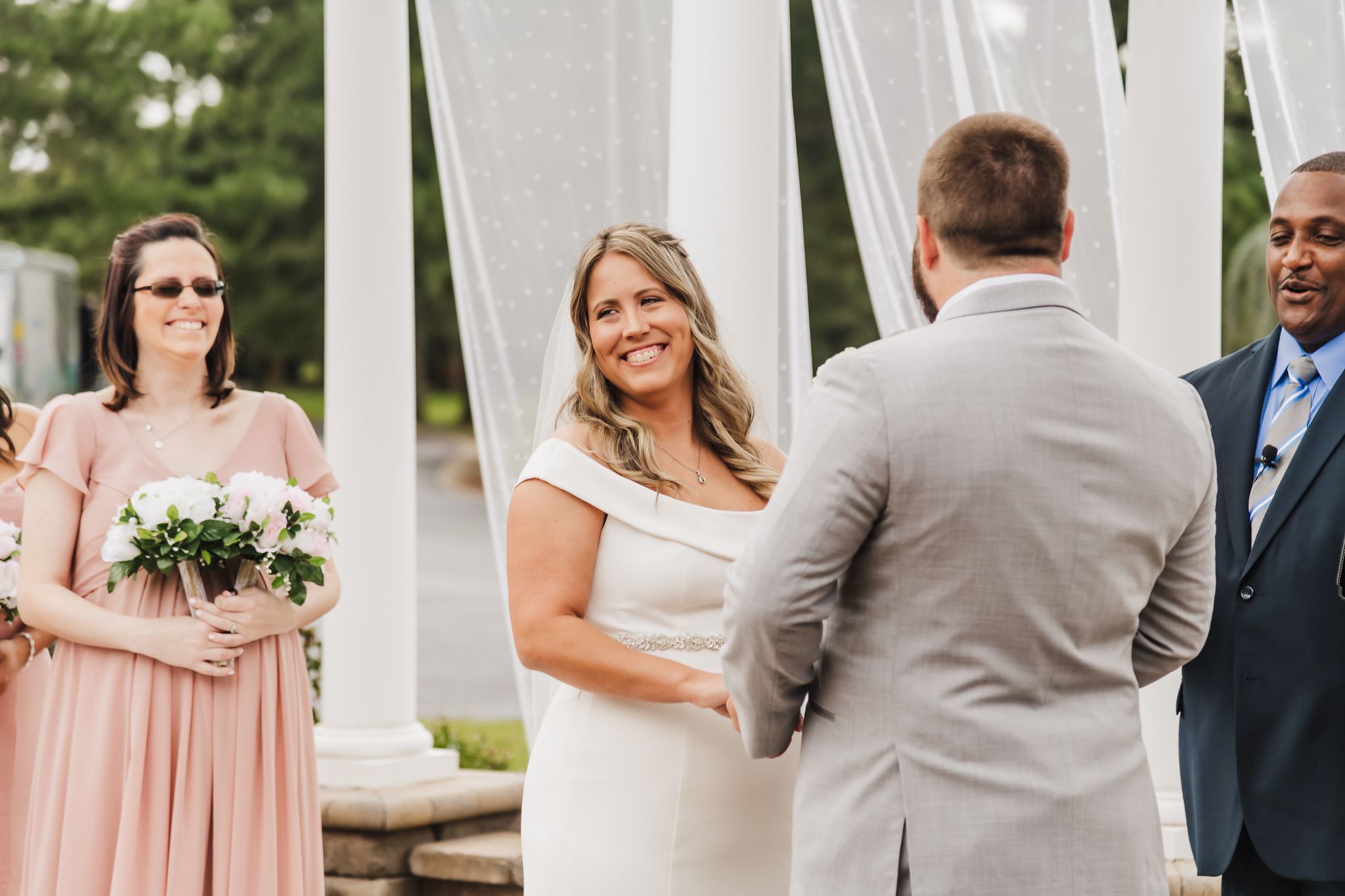 Wedding ceremony under white pergola with wedding party in attendance.