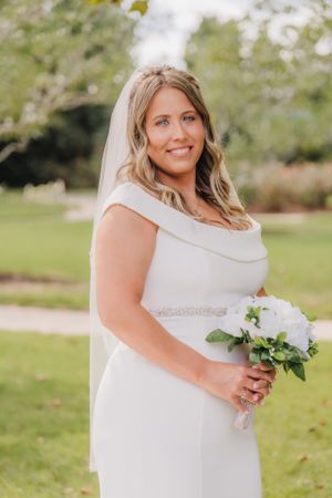 Bride holds white bouquet while posing in off-shoulder wedding dress outdoors.