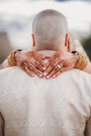 Henna-decorated hands embrace groom from behind wearing cream sherwani.