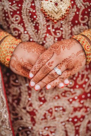 Close-up of bridal hands adorned with henna, bangles, and engagement ring on red lehenga.