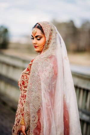 Bride in red lehenga and sheer veil poses elegantly against outdoor backdrop.