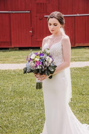 Beautiful bride in a lace white dress poses with a stunning purple and pink bouquet against a red barn backdrop.