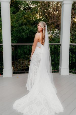 A bride in a fitted lace gown with long veil poses against white columns on a deck.