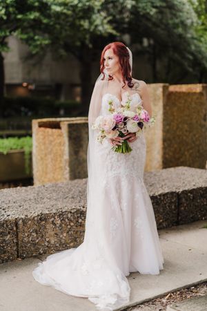 Bride in lace wedding dress holds pink and white bouquet while standing on stone steps.