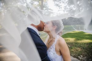 A dreamy wedding kiss captured through flowing veil with sunlit sprinklers in background.