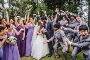 Wedding party celebrates with energetic jumping pose outdoors, bridesmaids in purple dresses and groomsmen in gray suits.