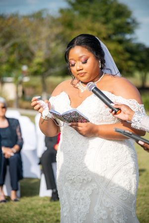 Bride in white lace dress reads from paper while holding microphone during outdoor ceremony.