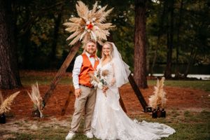 Wedding couple poses under dramatic pampas grass and floral arch in rustic outdoor setting.