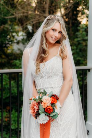 A bride in a lace gown holds a coral and white bouquet while posing on an outdoor deck.