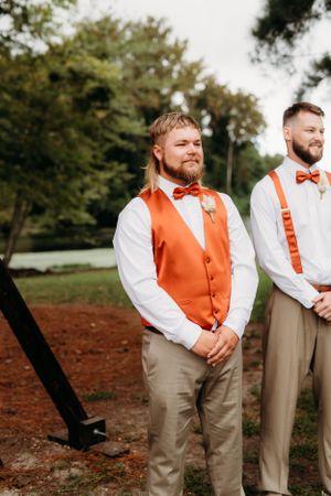 Groomsmen in orange vests and bowties stand during outdoor autumn wedding ceremony.