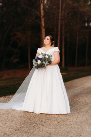 An elegant bride in an off-shoulder gown holds a blue and white bouquet on a garden path.