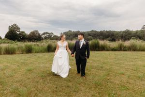 Couple walks hand in hand across grassy field in formal wedding attire.