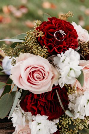 Wedding bouquet with deep red and blush pink roses, white flowers, greenery and engagement ring nestled in petals.