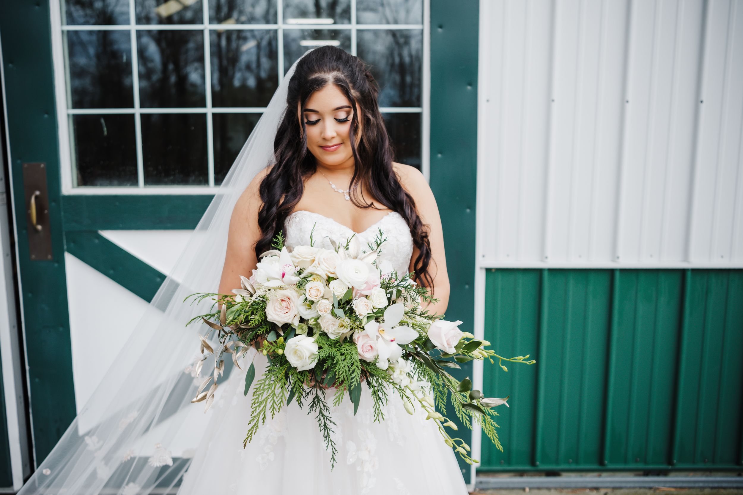 A close-up portrait of a bride holding a lush white and green bouquet against a rustic barn backdrop.