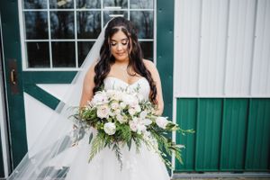 A close-up portrait of a bride holding a lush white and green bouquet against a rustic barn backdrop.