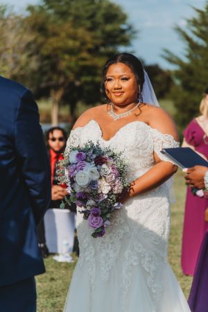 Bride in an off-shoulder lace gown holds a purple and white bouquet during an outdoor wedding ceremony.