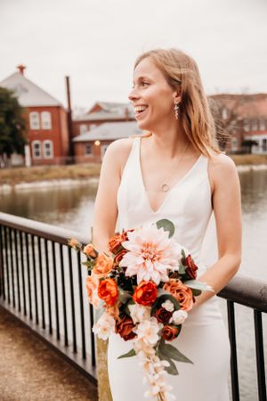 A bride in white dress smiles while holding coral and pink bouquet by waterfront railing.