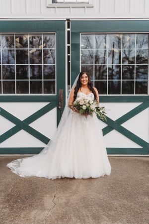 A bride poses with her bouquet in front of green and white barn doors wearing a stunning ballgown wedding dress.