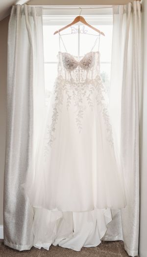 Elegant white wedding dress with intricate lace details hanging on wooden hanger against curtained window.