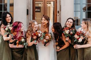 Bridesmaids in olive dresses laugh joyfully with their bouquets of pink and red flowers.