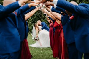 Wedding party in blue and burgundy attire forming arch with raised arms outdoors.