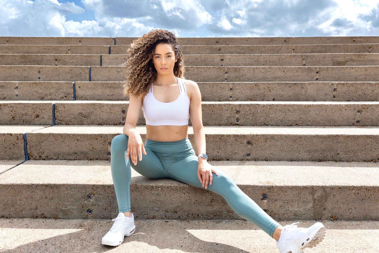Athletic woman in workout gear stretches on concrete steps under cloudy sky.