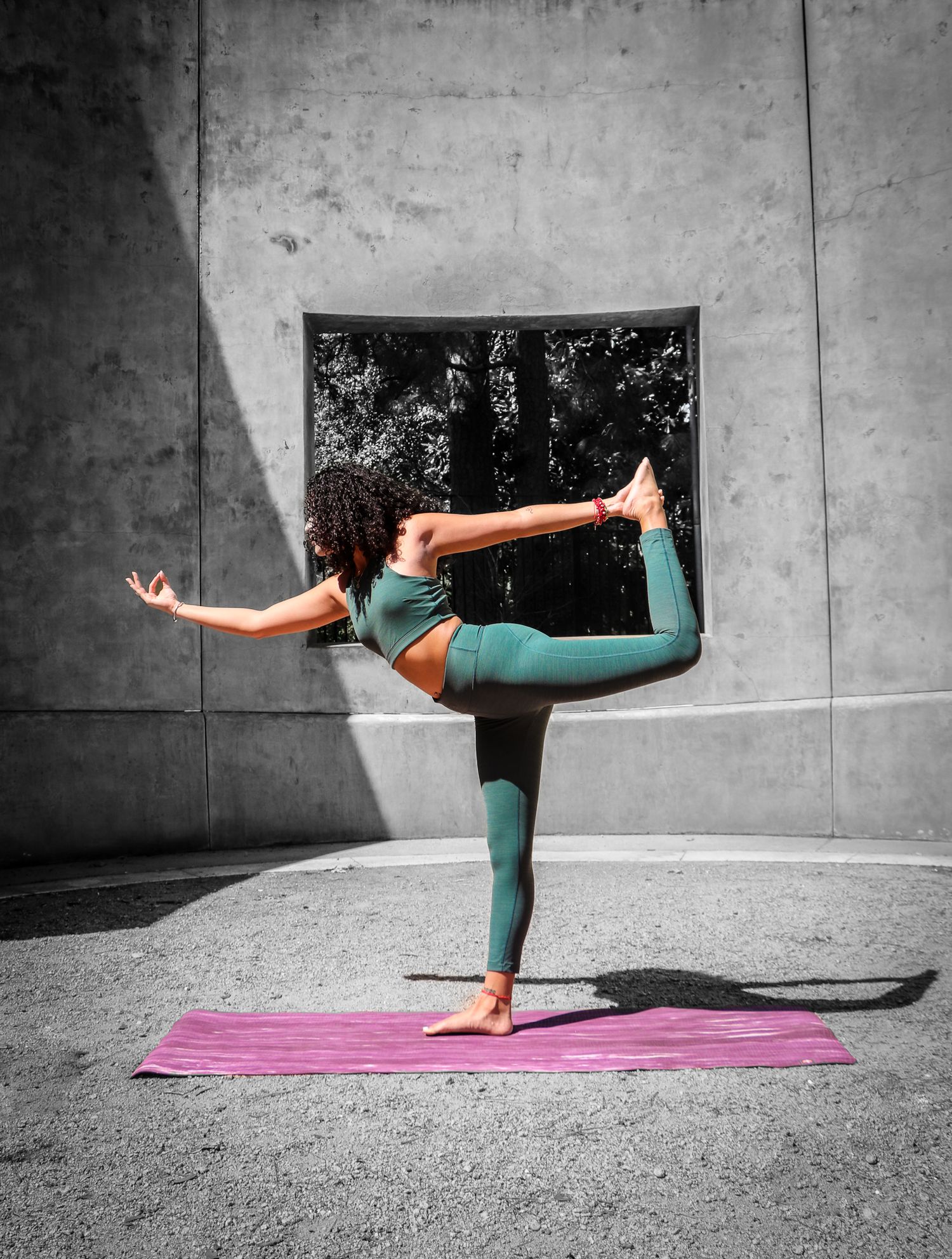 A yoga practitioner in teal activewear performs a standing pose on a pink mat against a gray wall.