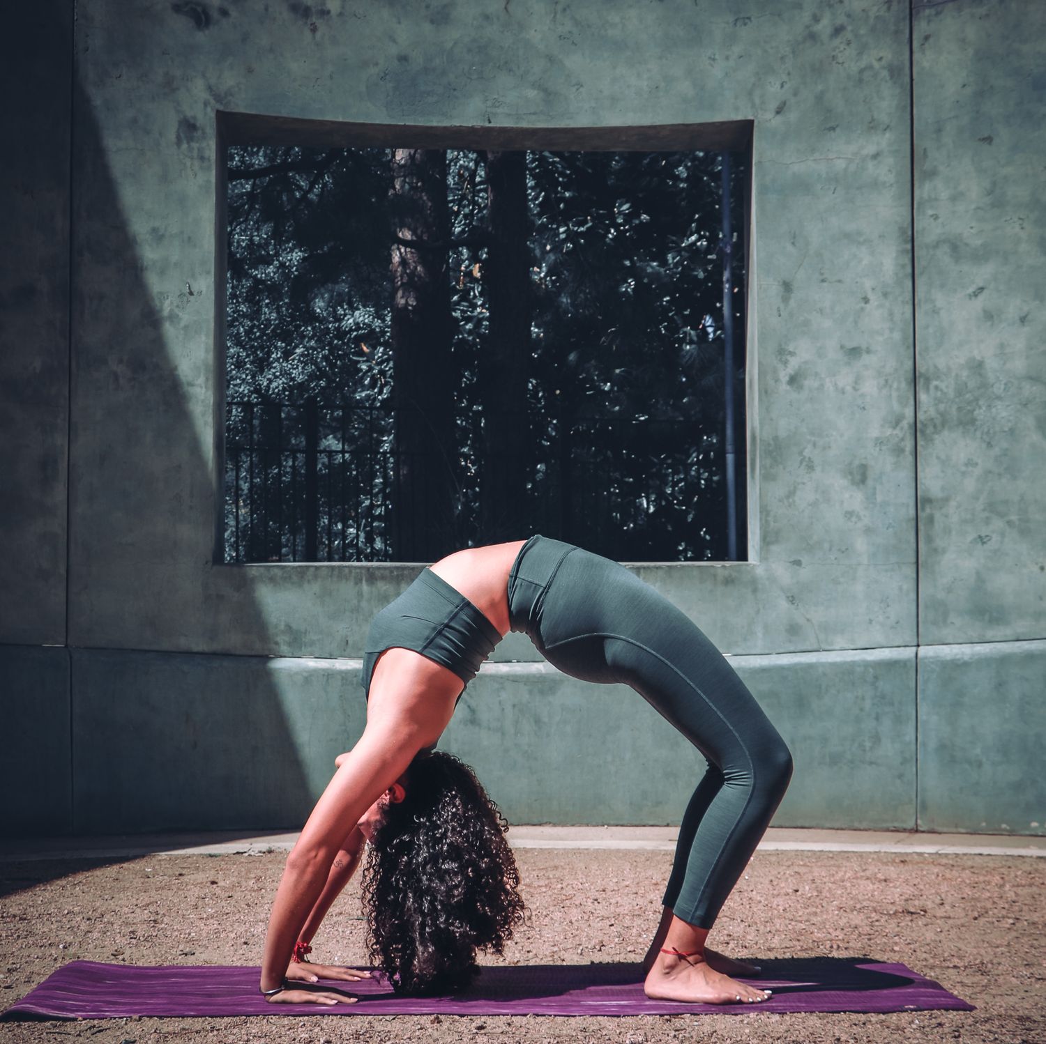 A yogi performs a wheel pose on a purple mat against an industrial concrete wall with window.