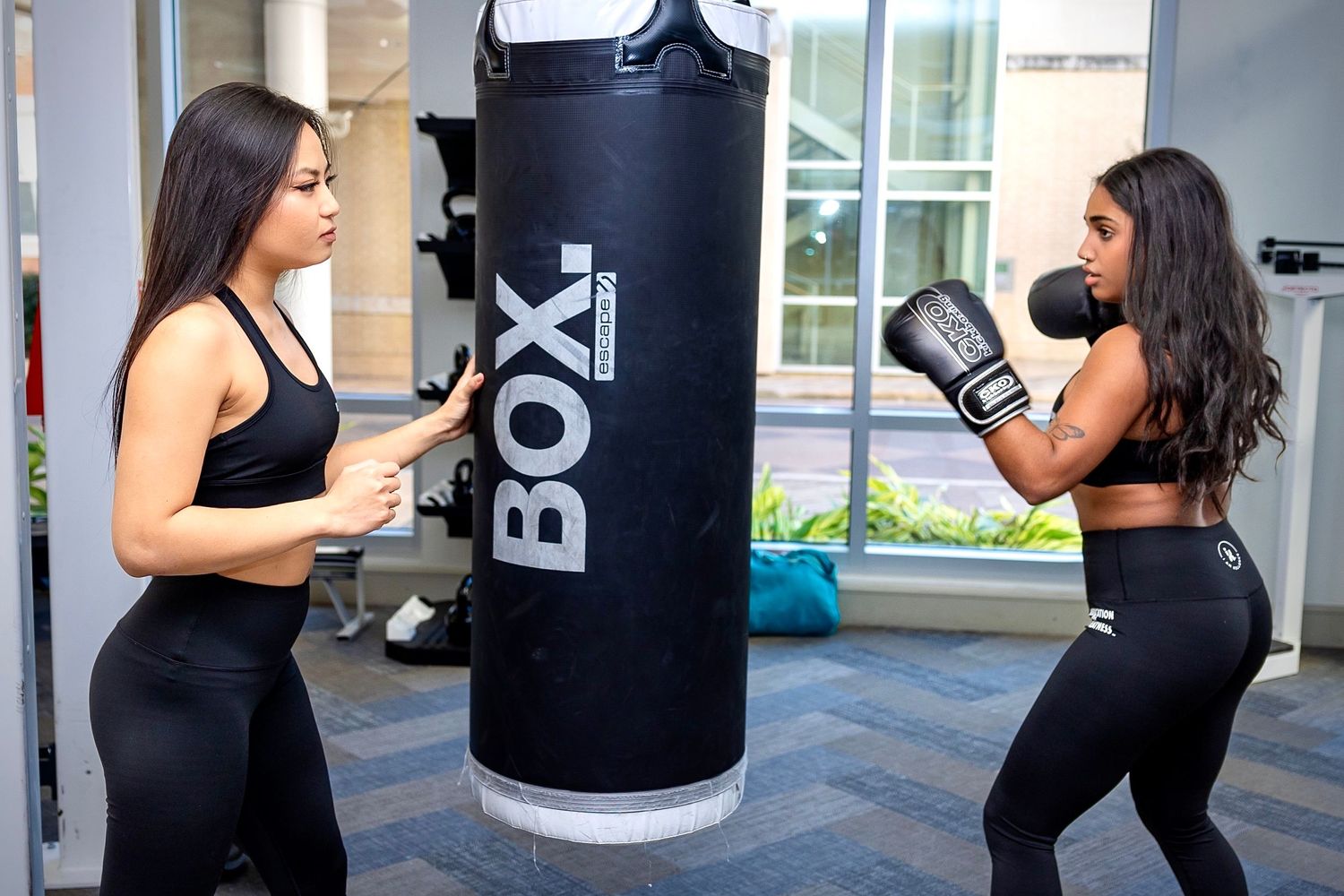 Two people practicing boxing moves with a heavy punching bag in an indoor gym setting.