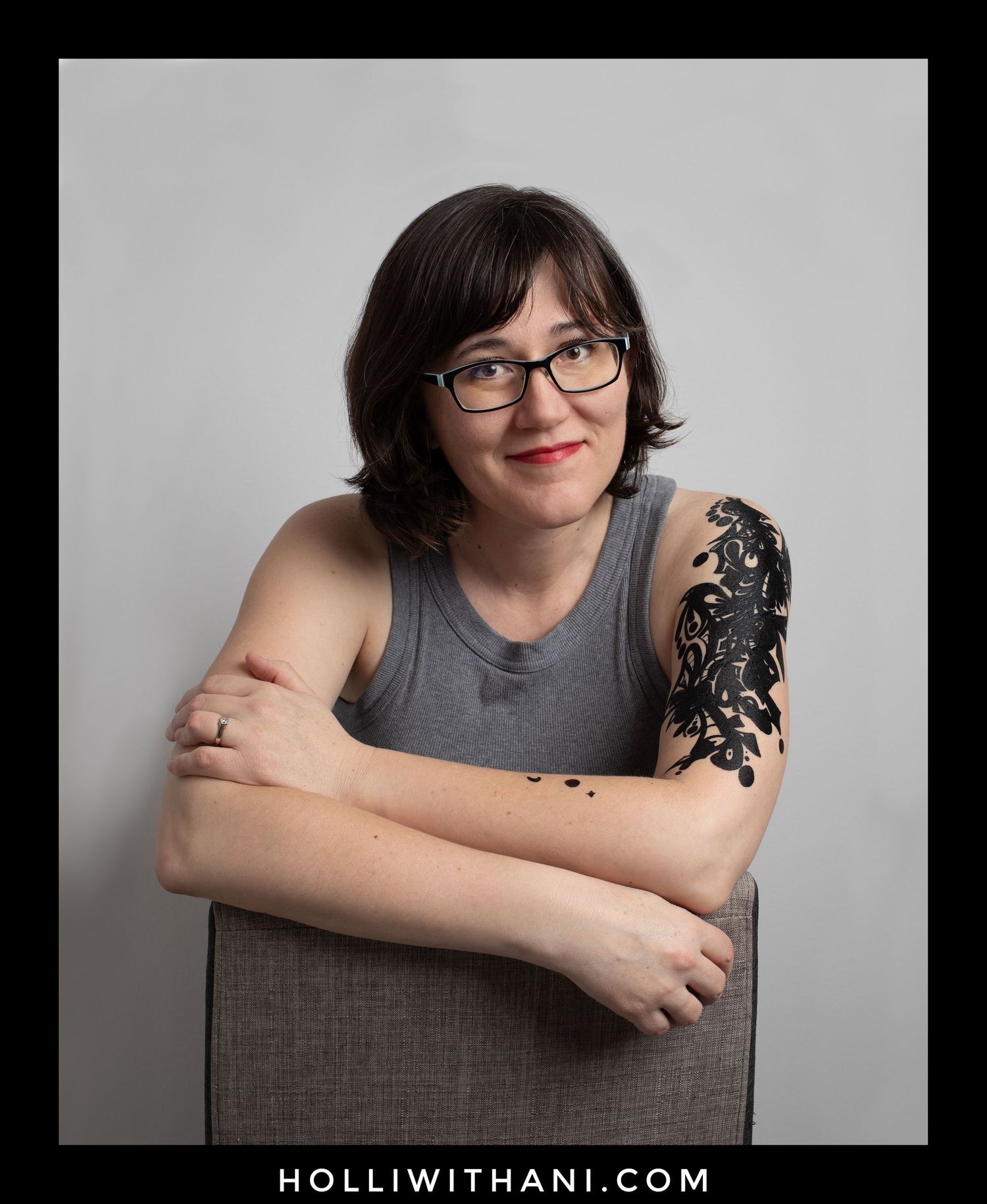 Portrait of Holli Margell sitting facing the camera with a sleeveless gray shirt showing arms tattoos