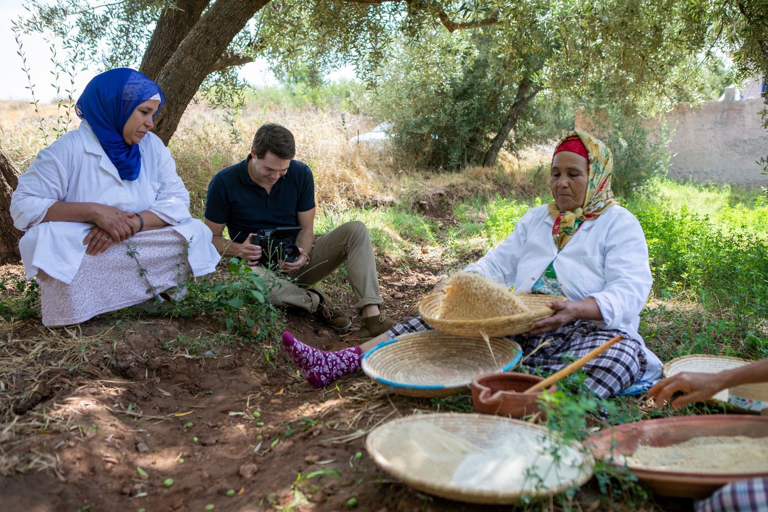 Traditional food preparation scene under an olive tree with woven baskets.