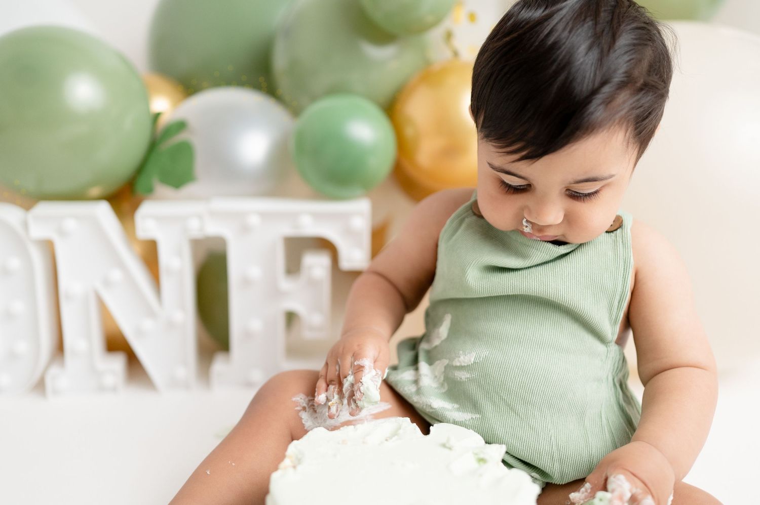 Baby in mint green romper enjoying cake smash photoshoot with green and gold balloon decor.
