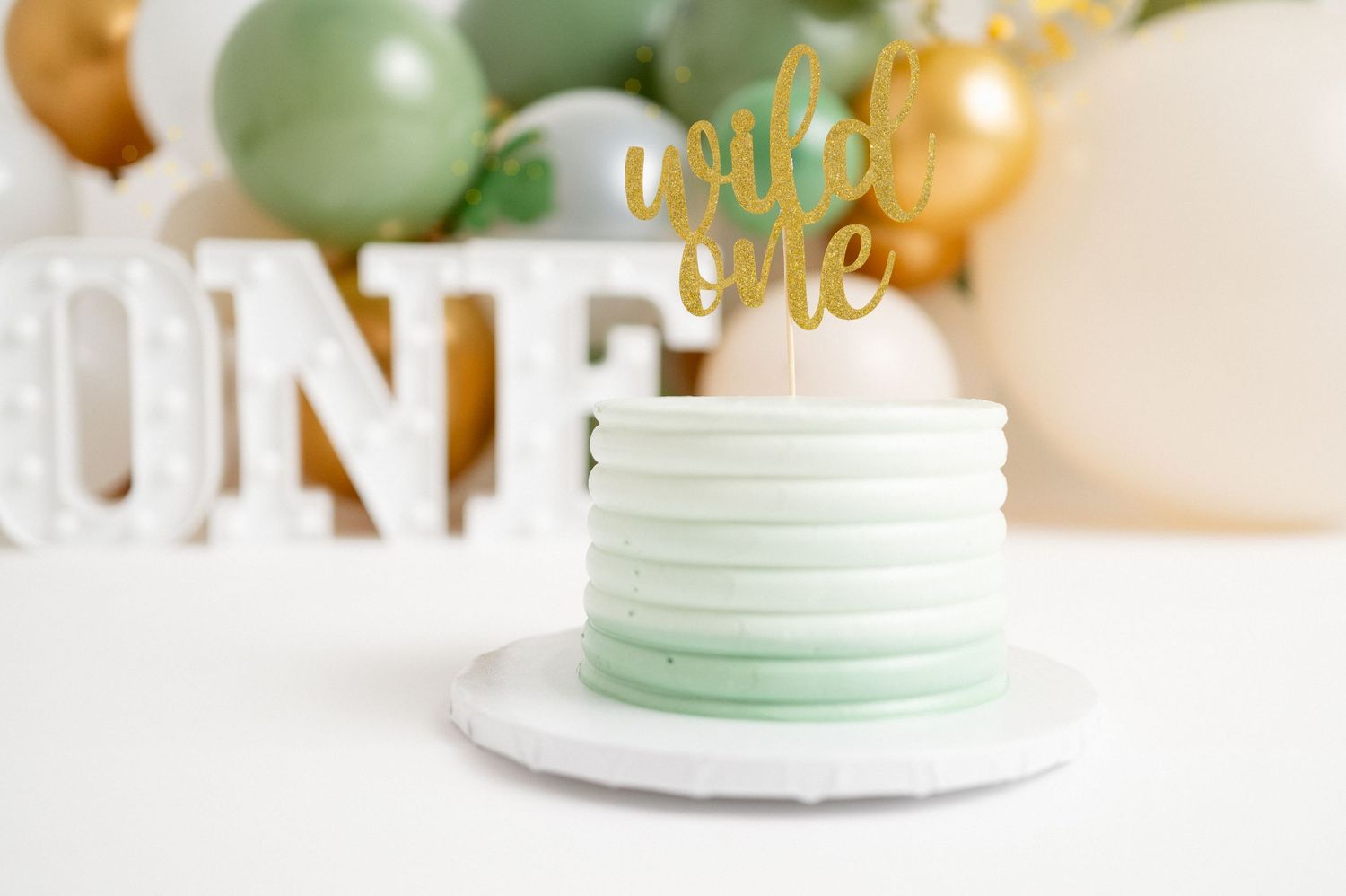 Mint green birthday cake with gold topper surrounded by balloon decorations.