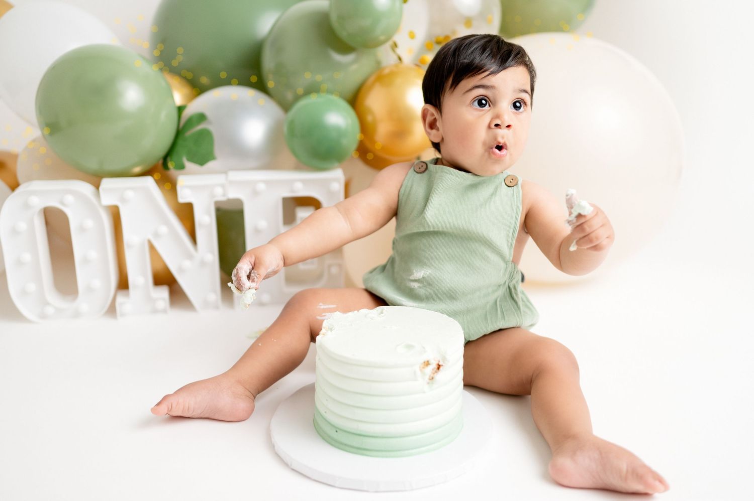 Baby in mint green outfit sitting next to white birthday cake with balloon decorations.